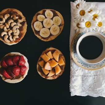 A selection of four small round snacks is arranged on a dark surface, each topped with different ingredients: cashews, banana slices, strawberries, and diced cantaloupe. To the right, a blue and white floral-patterned cup filled with black coffee sits on a white embroidered napkin adorned with fresh daisies.