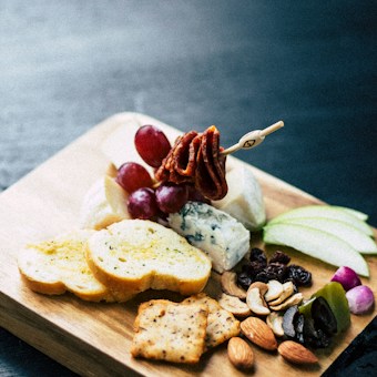 A wooden board displays a variety of snack items including sliced bread, green apple slices, grapes, various types of cheese, assorted nuts, olives, dried fruits, and crackers. A skewer with meat or vegetables is also visible, adding to the presentation.