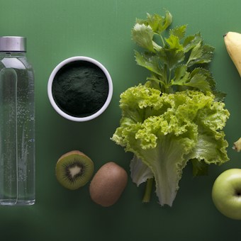 A neatly arranged assortment of healthy foods sits on a green background, including a clear water bottle, a bowl of dark green powder, leafy celery and lettuce, a banana, sliced kiwi, and a green apple.