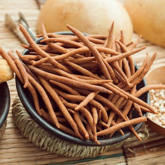 A collection of assorted snacks displayed in various bowls, including long, brown, finger-like snacks and round, golden, crispy balls. The arrangement is set on a textured, woven mat with additional small bowls containing seeds or grains. The background shows a soft focus with warm, earthy tones.
