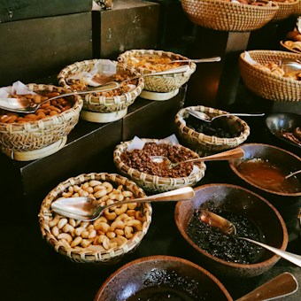 A variety of nuts, seeds, and dried fruits are displayed in woven baskets and bowls on a dark surface. Spoons are placed in each container for serving. The arrangement is visually appealing with an assortment of textures and shapes.