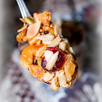 A close-up of a spoonful of mixed nuts, dried fruits, and grains. The mix includes almonds, cashews, raisins, and seeds. The background is blurred, drawing focus to the spoon's contents.