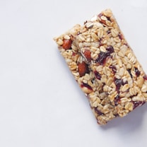 Two granola bars made from oats, nuts, and dried fruits are placed on a light gray background. The texture of the bars appears crunchy with visible almonds and cranberries.