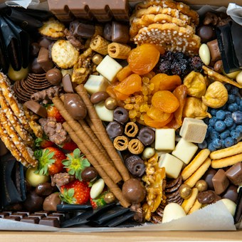 A box filled with a variety of snacks, including chocolates, dried fruits, nuts, fresh strawberries, wafers, and pretzels. The assortment is colorful and arranged neatly, featuring different textures and shapes.
