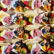 A collection of small paper trays filled with an assortment of snacks including fresh strawberries, raspberries, blueberries, blackberries, green grapes, almonds, dried apricots, crackers, slices of cheese, and rolled salami. The arrangement is colorful and appealing, presenting a variety of textures and flavors.