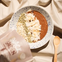 A bowl of smoothie topped with various ingredients like oats, coconut flakes, and seeds is placed on a patterned fabric. A wooden spoon lies next to the bowl, and a packet labeled 'Snack Garden' is partially visible on the side.