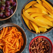 An arrangement of five bowls containing different snacks: red grapes in a wooden bowl, potato chips in a blue bowl, sliced mangoes in a brown bowl, cheese flavored snacks in a dark brown bowl, and a mix of nuts in a red bowl.