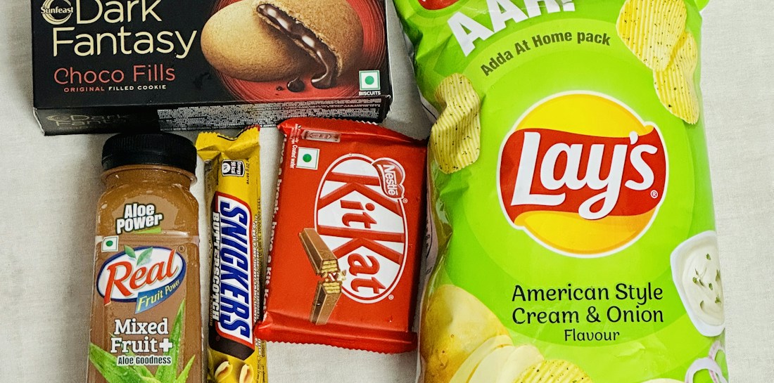 An assortment of packaged snack items including a bottle of mixed fruit juice, a Snickers bar, a Kit Kat bar, a box of Dark Fantasy Choco Fills biscuits, and a package of Lay's American Style Cream & Onion flavored chips. The items are laid out on a plain surface.